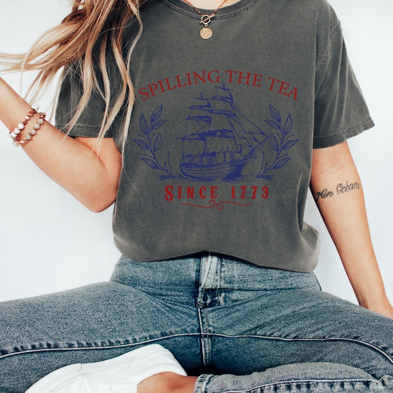 May include: Dark gray t-shirt with the text "Spilling the Tea Since 1773" above a blue illustration of a tall ship. The shirt is worn with blue jeans.
