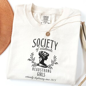 May include: A white t-shirt with a black graphic that says "Society of Obstinate Headstrong Girls seriously displeasing since 1813". The graphic features a silhouette of a woman with her hair up in a bun.
