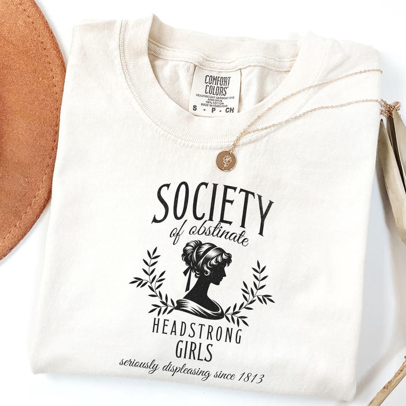 May include: A white t-shirt with a black graphic that says "Society of Obstinate Headstrong Girls seriously displeasing since 1813". The graphic features a silhouette of a woman with her hair up in a bun.