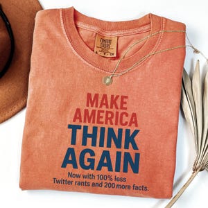 May include: A coral-coloured t-shirt with the text "Make America Think Again" in blue and red lettering. The text "Now with 100% less Twitter rants and 200 more facts." is printed below the main text.