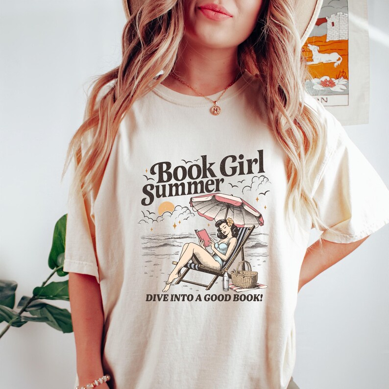 May include: Cream-colored t-shirt with a graphic that reads "Book Girl Summer." The graphic depicts a woman reading on a beach under an umbrella. The text "Dive into a good book!" is also included.