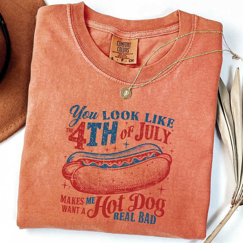 以下が含まれることがあります： 「You look like the 4th of July. Makes me want a Hot Dog real bad.」というテキストと、ビンテージのホットドッグのイラストが描かれた赤、白、青のグラフィックTシャツです。