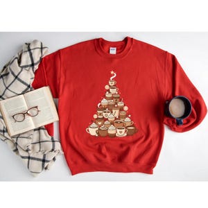 May include: A red sweatshirt features a Christmas tree design made of various coffee cups and mugs. The design includes different types of coffee drinks, such as lattes and cappuccinos. A star tops the tree. A book, glasses, and a mug are also in the image.