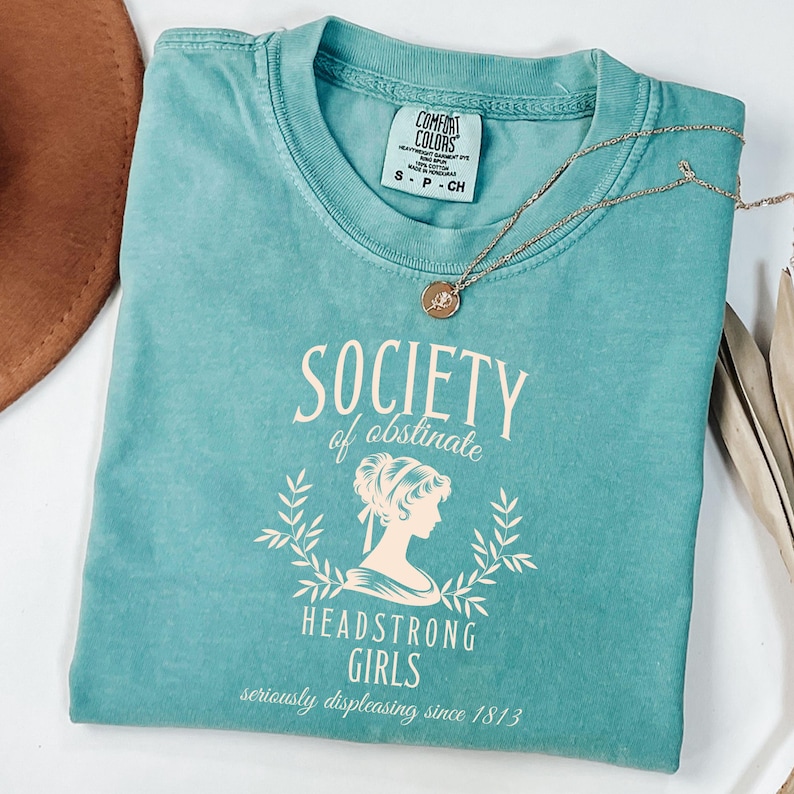 May include: Teal blue t-shirt with a vintage-style graphic that reads "Society of Obstinate Headstrong Girls seriously displeasing since 1813".
