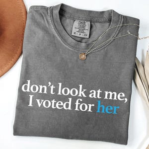 Peut inclure: T-shirt gris avec le texte "ne me regarde pas, j'ai vot&eacute; pour elle" en blanc avec le mot "elle" en bleu.