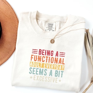 May include: A beige t-shirt with a vintage-style graphic that reads "Being a Functional Adult Everyday Seems a Bit Excessive". The graphic is in red, orange, yellow, green, and blue.