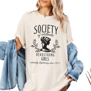 May include: A white t-shirt with black text that reads "Society of obstinate Headstrong Girls seriously displeasing since 1813". The text is surrounded by a black silhouette of a woman with her hair up and a floral design.