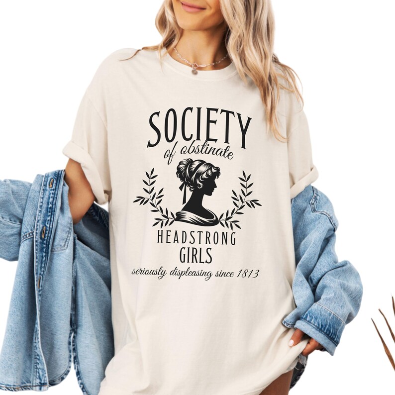 May include: A white t-shirt with black text that reads "Society of obstinate Headstrong Girls seriously displeasing since 1813". The text is surrounded by a black silhouette of a woman with her hair up and a floral design.