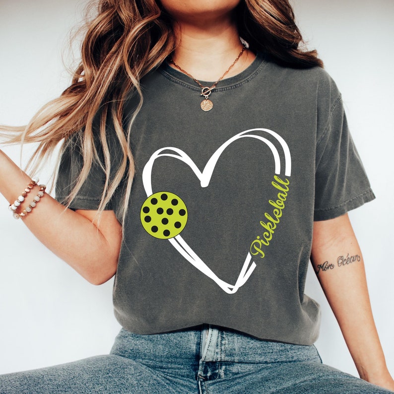 May include: A gray t-shirt with a white heart design featuring a green and black pickleball. The text "Pickleball" is written inside the heart.