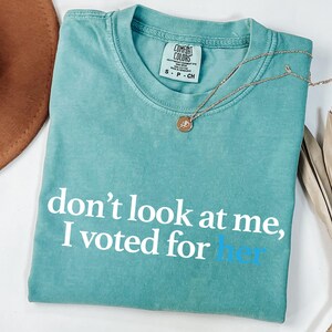 Peut inclure: Un t-shirt bleu turquoise avec le texte "ne me regarde pas, j'ai vot&eacute; pour elle" en blanc avec le mot "elle" en bleu.
