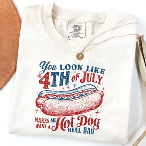 以下が含まれることがあります： 「You look like the 4th of July. Makes me want a Hot Dog real bad.」という赤、白、青のグラフィックがプリントされた白いTシャツ。グラフィックには、マスタードとケチャップをかけたホットドッグが描かれている。
