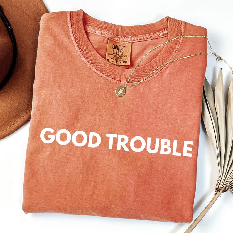 May include: A coral-colored t-shirt with the text "GOOD TROUBLE" printed in white. The shirt is made of a soft, comfortable fabric.