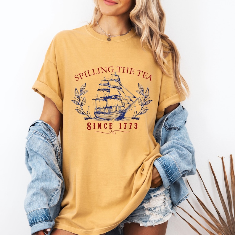 May include: Mustard yellow t-shirt with the text "Spilling the Tea Since 1773" and a navy blue illustration of a tall ship. The design is framed by laurel branches. The shirt has rolled-up sleeves.