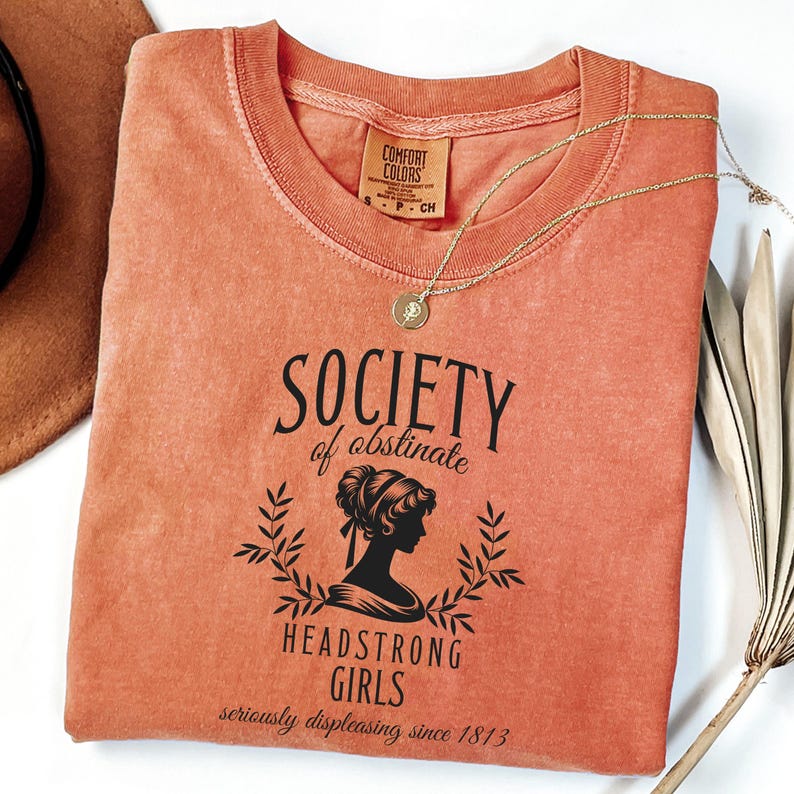 May include: A burnt orange t-shirt with a black graphic that reads "Society of Obstinate Headstrong Girls seriously displeasing since 1813". The graphic features a silhouette of a woman with her hair up in a bun.