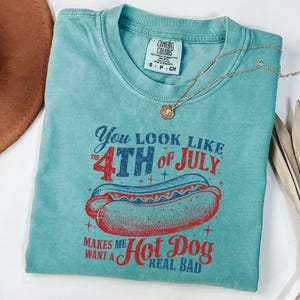以下が含まれることがあります： くすんだターコイズブルーのTシャツで、ホットドッグの赤、白、青のグラフィックが描かれており、"You look like the 4th of July Makes me want a Hot Dog Real Bad"というテキストが書かれています。