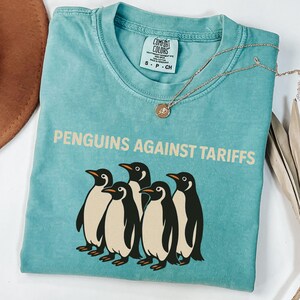 May include: A teal blue t-shirt with a vintage-style graphic of five penguins and the text "PENGUINS AGAINST TARIFFS".