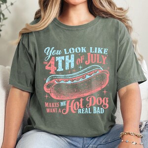以下が含まれることがあります： 「You look like the 4th of July makes me want a hot dog real bad」と書いてあるヴィンテージスタイルのグラフィックがプリントされた緑色のTシャツ。グラフィックには、星がついた赤、白、青のホットドッグが描かれている。