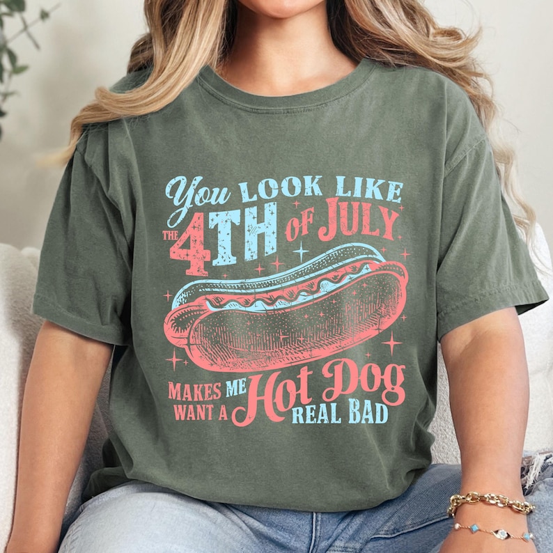 以下が含まれることがあります： 「You look like the 4th of July makes me want a hot dog real bad」と書いてあるヴィンテージスタイルのグラフィックがプリントされた緑色のTシャツ。グラフィックには、星がついた赤、白、青のホットドッグが描かれている。