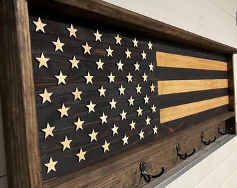 May include: A wooden wall-mounted coat rack with a distressed American flag design. The flag features a black background with gold stars and two horizontal stripes in natural wood tones. The rack has five black metal hooks.