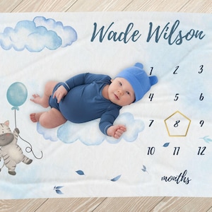 May include: A white baby blanket with a blue watercolour cloud design. The blanket has a number chart for tracking a baby's growth, with the name "Wade Wilson" at the top. A baby is lying on the blanket, and a cartoon zebra with a blue balloon is in the bottom left corner.