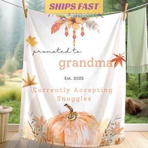 May include: White blanket with the text "promoted to grandma" and "Currently Accepting Snuggles." The design includes a watercolor pumpkin, autumn leaves, and floral accents. The blanket is hanging on a clothesline.