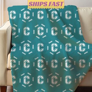 May include: Teal blanket with a repeating pattern of the letter "C" in white, with a baseball design inside the letter. The name "Connor" is printed around each letter. The blanket is draped over a beige sofa.