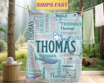 May include: A personalized baby blanket featuring a light blue background with various shark illustrations. The name "Thomas" is prominently displayed in large teal letters, with the name repeated in a pattern. The blanket is hanging on a clothesline.