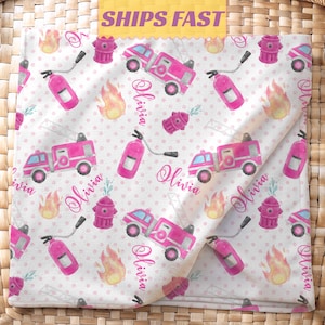 May include: A white blanket with a pink polka dot pattern featuring pink fire trucks, fire hydrants, flames, and fire extinguishers. The name "Olivia" is written in cursive. The blanket is folded on a woven basket.