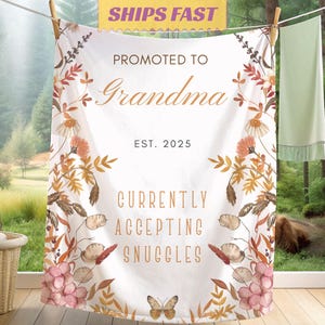 May include: A white blanket with floral and butterfly designs, hanging on a clothesline. Text reads "Promoted to Grandma EST. 2025" and "Currently Accepting Snuggles". The blanket has a rustic, autumnal theme.