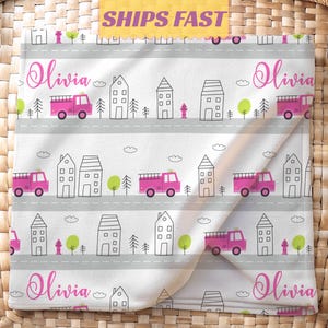May include: A white baby blanket with a repeating pattern of pink fire trucks, houses, trees, and the name "Olivia" in pink script. The blanket has gray roads and the text "SHIPS FAST" in a yellow banner.