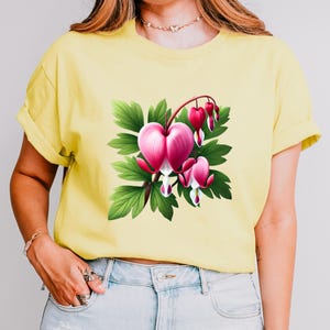 Op de afbeelding: Geel T-shirt met een gedetailleerde illustratie van een bloedend hart bloem. Het ontwerp toont een grote, roze hartvormige bloem, kleinere hartvormige bloemen en groene bladeren. Het shirt is gemaakt van zacht materiaal.