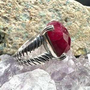 May include: A silver ring with a large, faceted, deep red gemstone. The ring's band has a twisted rope design. The gemstone is set in a prong setting. The ring is displayed on a bed of purple crystals.