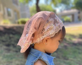 May include: A blush-colored head covering with floral lace trim. The delicate fabric drapes over the head, showcasing intricate embroidered floral patterns in shades of white and pale pink. The head covering is worn by a young child.