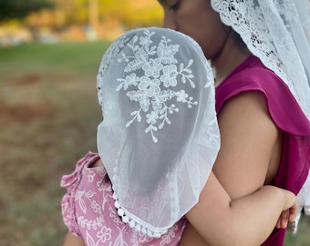 Può includere: Un velo di pizzo bianco con ricamo floreale e bordatura a pompon. Il velo è indossato da una persona che tiene un bambino.