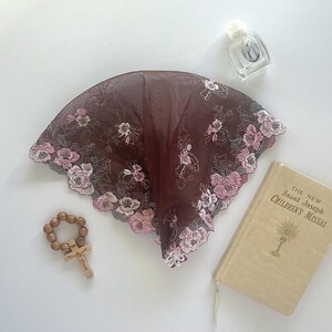 Może przedstawiać: Burgundowa mantilla z koronki z haftowanymi r&oacute;żowymi i białymi kwiatami. Mantilla leży na białym tle z drewnianym r&oacute;żańcem i małą książką zatytułowaną "The New Saint Joseph Children's Missal".