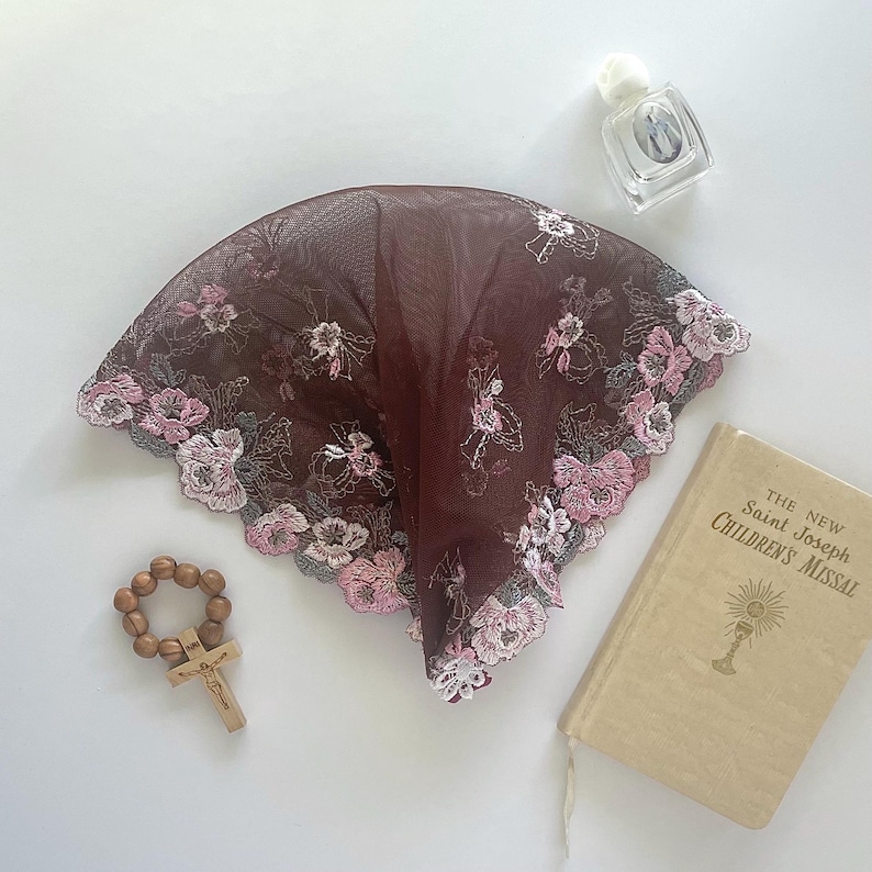 Może przedstawiać: Burgundowa mantilla z koronki z haftowanymi r&oacute;żowymi i białymi kwiatami. Mantilla leży na białym tle z drewnianym r&oacute;żańcem i małą książką zatytułowaną "The New Saint Joseph Children's Missal".