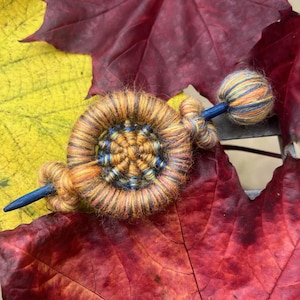May include: A handmade hair pin with a woven, circular design in shades of orange, yellow, and blue. The pin is set against a backdrop of vibrant red and yellow autumn leaves, creating a warm, seasonal aesthetic.