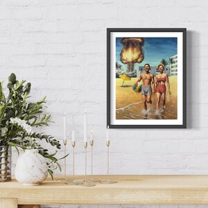 May include: Framed art print depicting a couple on a beach, seemingly oblivious to a mushroom cloud in the background. The man holds a beach ball, and the woman wears a red swimsuit. The artwork has a retro, vintage aesthetic.