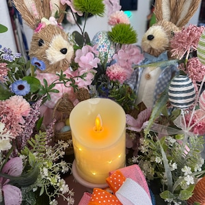 Może przedstawiać: Wiosenny element dekoracyjny z dwoma figurkami królików, różowymi i niebieskimi kwiatami, zapalonym świecą i biało-różowo-pomarańczową wstążką.