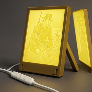 Op de afbeelding: Een heldergele LED-fotolijst met een gedetailleerde afbeelding van een persoon met een hert. De lijst heeft een ingebouwde standaard en een wit snoer met een aan/uit-schakelaar. Een tweede lijst met een silhouet is zichtbaar.