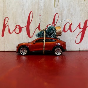 May include: A miniature orange car ornament with a small green Christmas tree tied to the roof. The ornament has silver wheels and a silver hook for hanging. The word "holiday" is visible in the background.