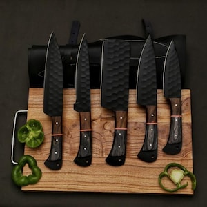 以下が含まれることがあります： 木製のまな板の上に、木製のハンドルが付いた黒いキッチンナイフ6本セット。ナイフは、ハンマーで叩いたような質感の仕上げになっています。