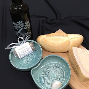 May include: A teal ceramic dipping bowl set with a garlic clove, bread, cheese, and olive oil. The set includes a bottle of olive oil, a loaf of bread, a block of cheese, and a small packet of herbs. The dipping bowl has a spiral design.