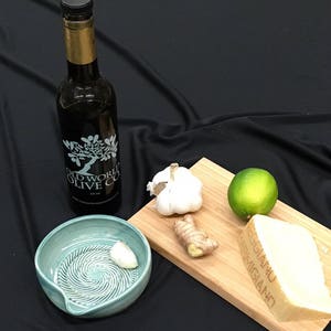 May include: A dark glass bottle of olive oil with a gold neck, a light blue ceramic bowl, and a wooden cutting board with ingredients like garlic, ginger, lime, and Parmesan cheese. The olive oil bottle has the text "Old World Olive Co."