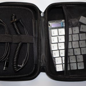Könnte beinhalten: Eine schwarze Tragetasche mit einem Reißverschluss. Die Tasche ist geöffnet und enthält eine kleine weiße Tastatur, ein schwarzes Kabel und ein schwarzes Spiralkabel. Die Tastatur hat Tasten mit Zahlen und Buchstaben beschriftet, darunter "1", "2", "3", "4", "5", "T", "R", "E", "W", "M" und "1".