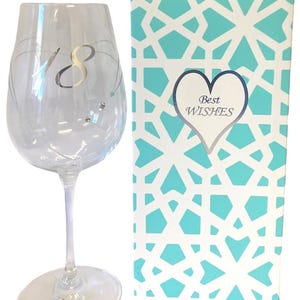 Puede incluir: Una copa de vino de vidrio transparente con un número 18 plateado y diamantes de imitación. La copa está en una caja de regalo con estampado turquesa y blanco con las palabras "Mejores deseos" en un corazón.