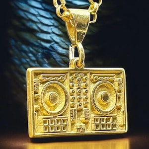 Może przedstawiać: Złoty łańcuszek z zawieszką w kształcie miksera DJ. Zawieszka ma dwa pokrętła, wyświetlacz i przyciski.