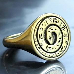 May include: A gold-toned signet ring featuring a circular face with an engraved spiral design. The engraving depicts a coiled snake surrounded by small celestial motifs including stars, crescent moons, and delicate leaf patterns.