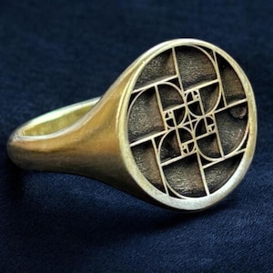 Könnte beinhalten: Ein goldener Siegelring mit einem runden Design, das ein geometrisches Muster aus sich überlappenden Kreisen zeigt, das der Fibonacci-Sequenz ähnelt.