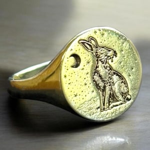 Puede incluir: Un anillo de sello en tono dorado con una cara circular grabada con un conejo detallado sentado bajo una luna creciente y un campo de pequeñas estrellas. La superficie metálica tiene un acabado pulido y la banda se estrecha hacia atrás.